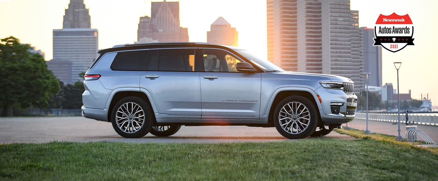 A profile view of a 2021 Jeep Grand Cherokee L Summit Reserve parked near a waterfront promenade with a cityscape at sunset in the background.