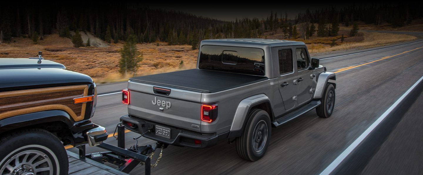 A 2022 Jeep Gladiator Overland being driven on a highway as it tows another vehicle on a flatbed trailer.